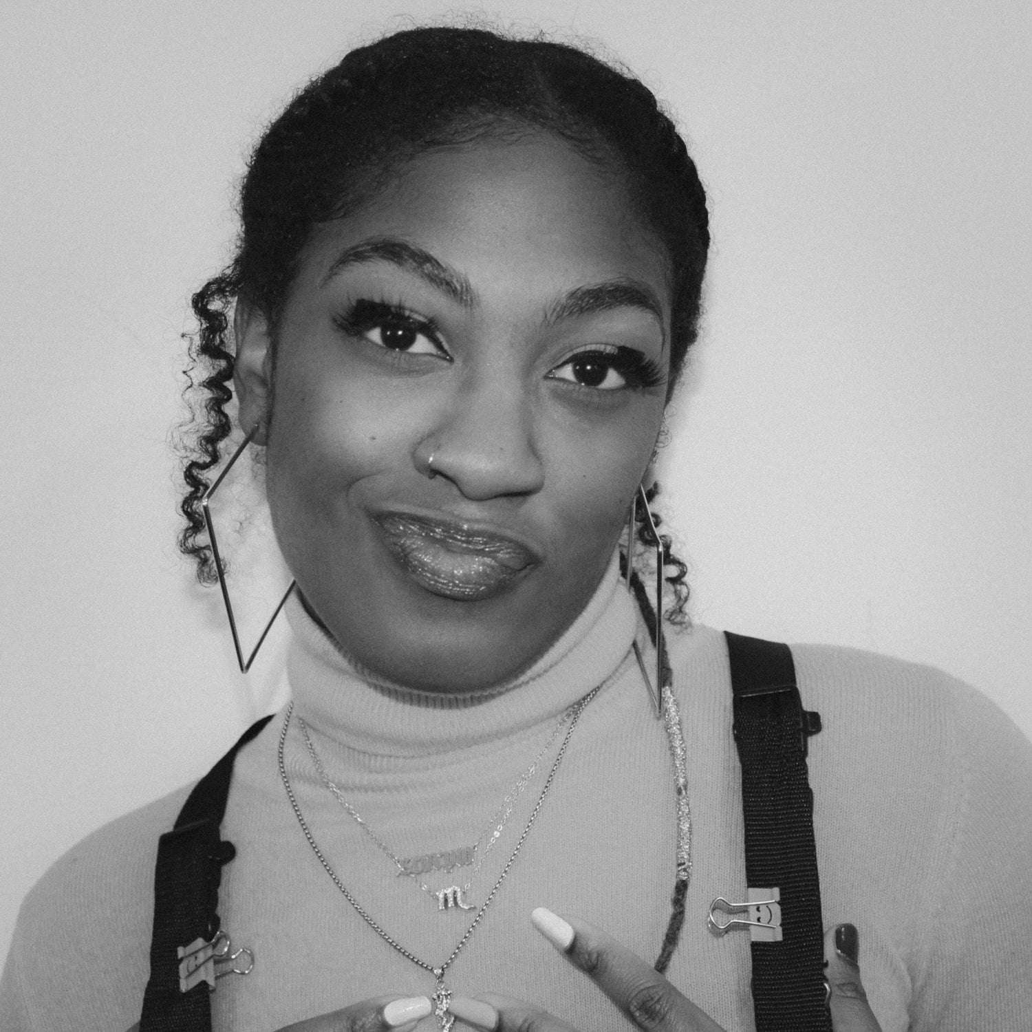 Artist, Laila P. FKATHECREATOR, posing and smirking. She is wearing a turtleneck and square earrings. Her hair is in slicked back and has her curls on the side of her face.